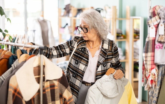 Senior woman client pre-examine fabric and style of product, is busy choosing down jacket or drap coat. Client enjoys shopping, clothing and accessories for whole family, seasonal sale