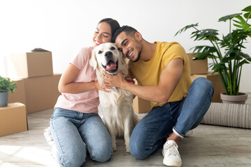 A young couple sits on the floor smiling while hugging their golden retriever. They are surrounded...