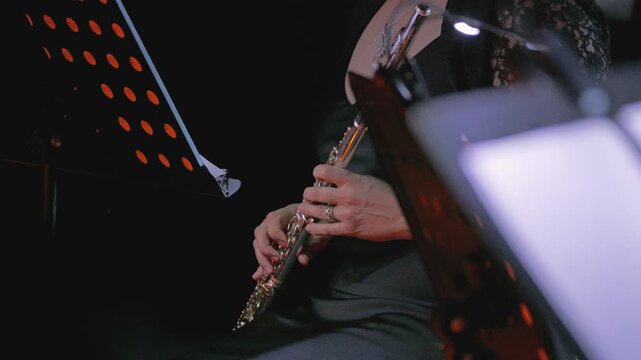 A close-up of a woman in a black dress holding a silver flute. The flute rests on her lap, and a music stand is partially visible nearby. The musician is ready to play at any moment