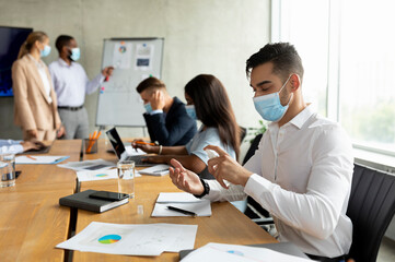 Arab male employee in a white shirt applies sanitizer on his hands while sitting at a desk in a contemporary office during a meeting with colleagues wearing medical masks.