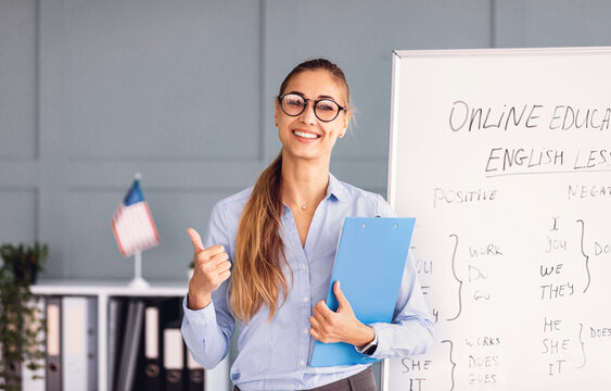 A confident teacher stands near a whiteboard in a bright classroom, sharing insights on online education while giving a thumbs up. The board displays notes about English lessons.