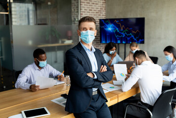 A company CeO, wearing a medical mask, stands confidently in a boardroom with coworkers. The team, diverse in background, collaborates on work during the Covid pandemic.