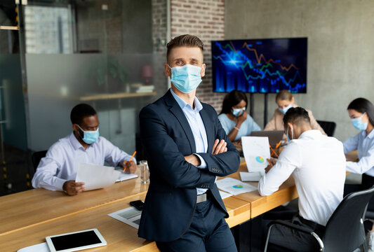 A company CeO, wearing a medical mask, stands confidently in a boardroom with coworkers. The team, diverse in background, collaborates on work during the Covid pandemic.