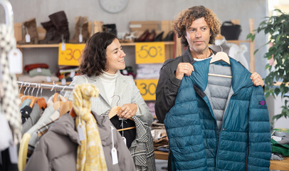 Happy european couple examine and choose new coat or down jacket for cold season in big modern store