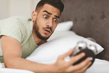 A young man with a surprised expression wakes up in bed, startled by the time on his alarm clock. He appears worried as he realizes he has overslept and is late for an important event.