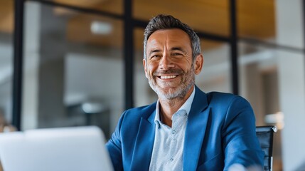 Naklejka premium A confident man with grey hair is seated at a desk in a sleek office. He is engaged with his laptop smiling warmly. Large windows allow natural light to fill the room.