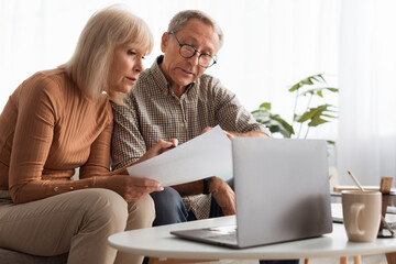 An older couple is focused on reviewing important documents together at home. They sit comfortably on a couch with a laptop nearby and natural light streaming in.