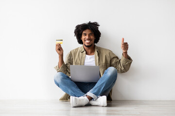 A cheerful Black man sits on the floor near a white wall, using a laptop and holding a credit card. He shows a thumbs up, recommending online banking for easy payments.