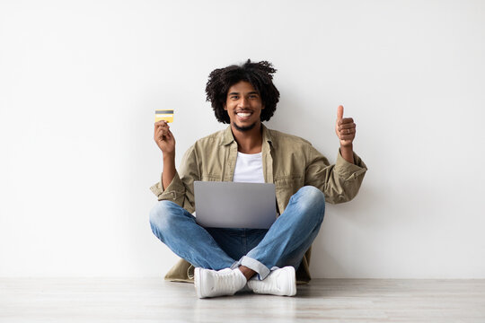 A cheerful Black man sits on the floor near a white wall, using a laptop and holding a credit card. He shows a thumbs up, recommending online banking for easy payments. - Powered by Adobe