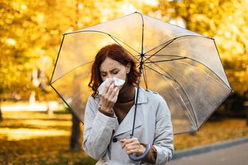 Naklejka premium A woman with red hair is standing in a bright autumn park, looking worried while holding a tissue in one hand and a clear umbrella in the other. Leaves fall around her.