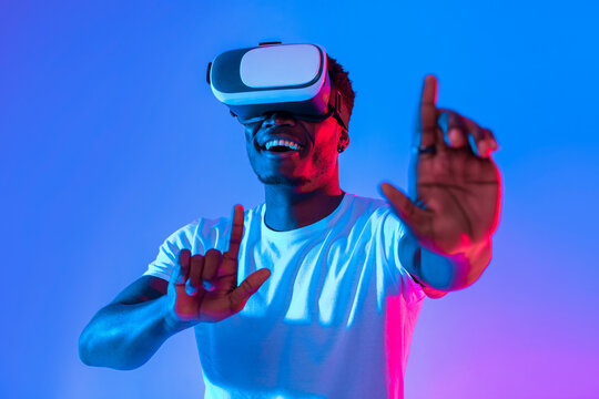 In a playful environment, a young man wears a virtual reality headset and smiles broadly. His hands are raised as he interacts with the virtual world, surrounded by bright blue and pink lights. - Powered by Adobe