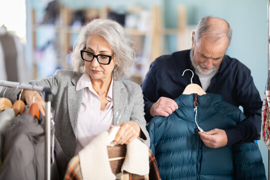 Senior wife with husband client pre-examine fabric and style of product, is busy choosing down jacket or drap coat. Client enjoys shopping, clothing and accessories for whole family, seasonal sale