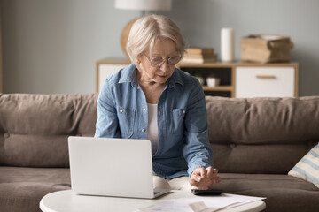 Smart retirement budgeting. Busy focused stylish older lady in glasses sit on sofa by laptop at home office count on calculator manage financial papers plan monthly expenses pay bills analyze receipts