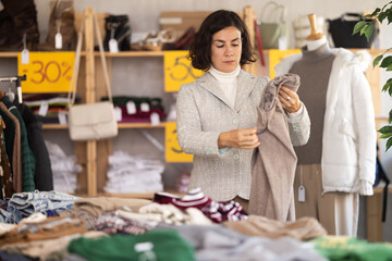European woman stands near the counter with warm winter clothes and chooses woolen pants for the cold season. Woman is shopping in the interior of a store