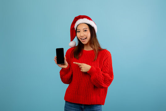 Christmas mobile offer. Happy asian lady in Santa hat pointing at smartphone with blank screen demonstrating free place for your advertisement, website or app design, blue background, mockup - Powered by Adobe