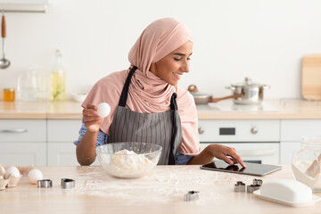 A woman wearing a headscarf is baking in a bright kitchen. She is holding an egg in one hand while checking a recipe on her tablet, surrounded by flour and baking tools.