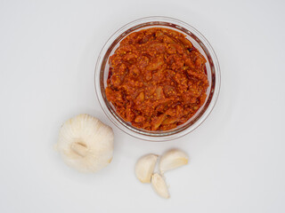 High-angle studio shot of Indian garlic pickle in a glass bowl with raw garlic cloves on a white background