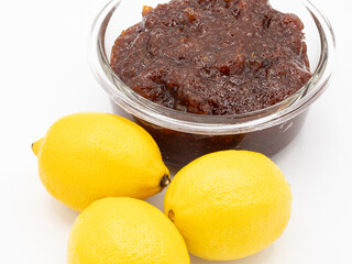 Indian lemon pickle in glass bowl with three fresh lemons against a white background