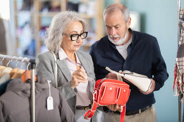 Mature couple spend time in shop and choose eco leather handbag. Changing wardrobe, shaping style of clothing. Designer clothes, clothing of world brands. Client elect between several goods options