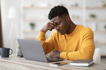Tired african american young man manager sitting at workdesk at office, working on laptop, touching his head, suffering from headache, experiencing financial crisis with business, copy space