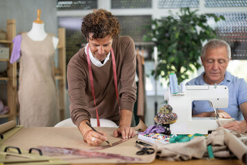 Fashion designer draws a sketch and a size grid on paper, next to a male tailor who works on a sewing machine. Men create a collection of clothes in a sewing workshop