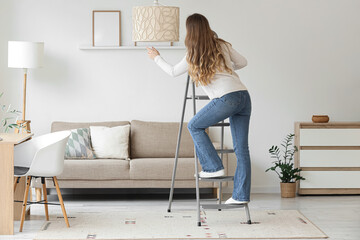 Young woman climbing stepladder near lamp in living room, back view