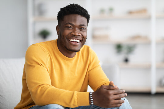 Portrait of successful millennial black guy in casual outfit sitting on couch in cozy living room and smiling at camera, posing alone at home, copy space. Millennials lifestyles concept