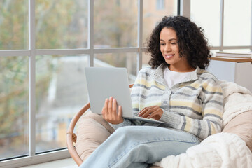 Young African-American woman working with laptop in armchair at home