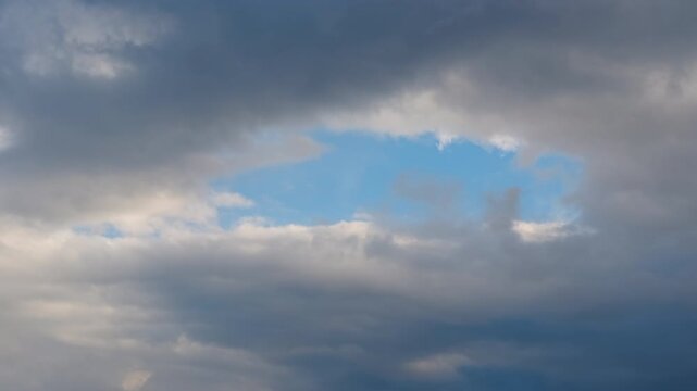 Clouds are slowly moving creating a hole in the sky revealing the blue. Slow-moving white and gray clouds drifting across sky, gradually parting to unveil bright blue atmospheric expanse behind