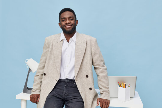 Confident smiling businessman in beige blazer and white shirt standing in modern office with desktop computer and accessories, looking at camera, indoor workplace, professional attire, casual style - Powered by Adobe