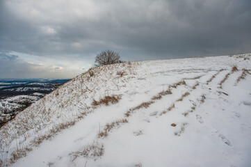 Dunkle Wolken &uuml;ber Schneelandschaft aufsteigender Wanderweg Gustav-Str&ouml;hmfeld-Weg und Eduard M&ouml;rike Weg am ehemaligen Vulkanschlot Jusiberg in Kohlberg vor grauem Himmel. mit kahlem Baum