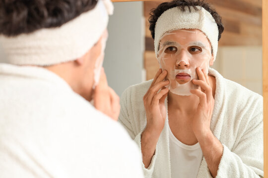 Young man applying facial sheet mask near mirror in bathroom