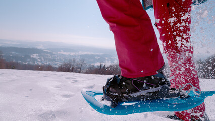 CLOSE UP, LOW ANGLE VIEW: Woman in pink pants breaking through fresh snow in snowshoes, sending crystals flying in the air on a sunny winter day. Scenic hike through the snowy hilly countryside.