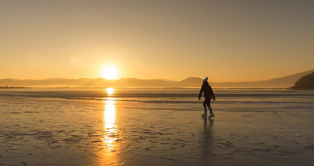 Fototapeta premium LENS FLARE, SILHOUETTE: Woman ice skating on frozen Lake Cerknica at sunrise, surrounded by golden morning light and soft outlines of the mountains in background. Outdoor sports in cold winter days.