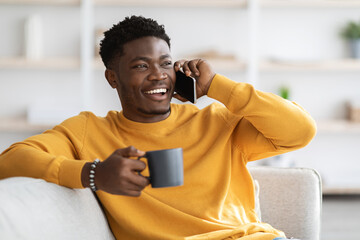 Cheerful black guy in yellow sitting on sofa, having phone conversation with friend and laughing, holding mug with coffee, looking at copy space. Handsome millennial black man spending weekend at home