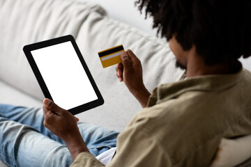 A black man lounges on a couch at home, using a digital tablet and credit card to make online purchases. He appears focused and relaxed while enjoying e-commerce at his leisure.