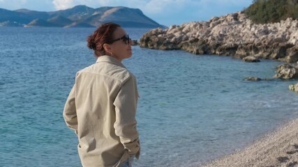 Woman enjoying a peaceful walk along a serene beach coastline. Woman strolls along a tranquil beach, taking in the calm ocean views and rocky shoreline. Perfect depiction of relaxation and solitude