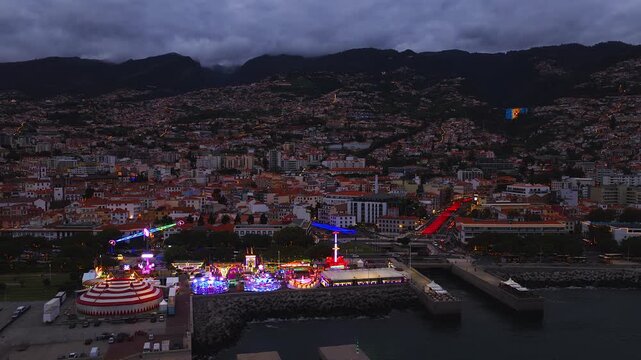 Aerial view of Funchal, Madeira, Portugal at dusk. Marina lights, a red and white circus tent, rides, traffic, and a large Christmas tree glow against dark hillsides.