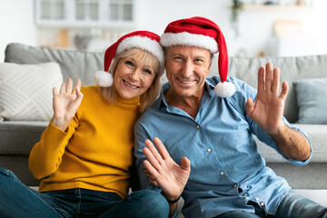 A joyful couple sits on a cozy couch, wearing Santa hats and smiling cheerfully. They wave at the camera, creating a warm holiday atmosphere filled with love and togetherness.