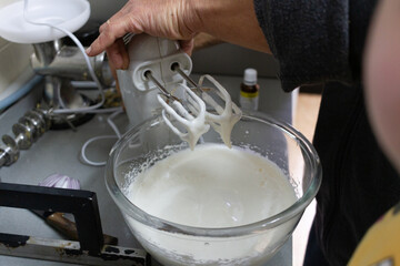 Close up of an adult hand using an electric mixer to whip thick cream in a glass bowl, while a curious child looks on, helping to bake dessert in a small family kitchen, together on a quiet weekend.