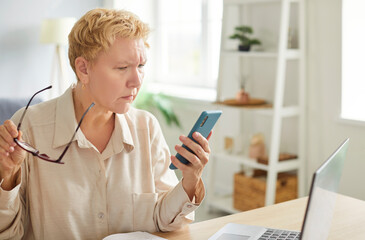 Focused woman with short blond hair sitting at desk with laptop, holding eyeglasses, looking at her...