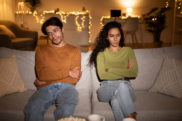 In the evening, an unhappy couple sits apart on their sofa, arms crossed. They look distant after a...