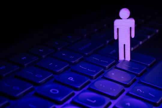 Wooden figure stands on a laptop keyboard illuminated by purple light, symbolizing technology and digital interaction in modern life