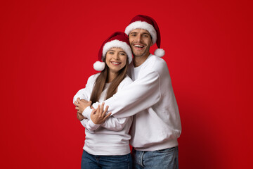 A cheerful couple embraces in front of a bright red background, both wearing warm sweaters and...