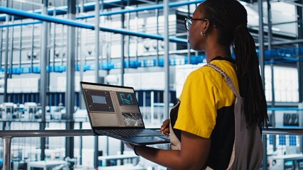 Employee using laptop in smart factory, collecting and analyzing data. African american woman using tracking software in industry 4.0 industrial plant equipped with advanced sensors, camera A