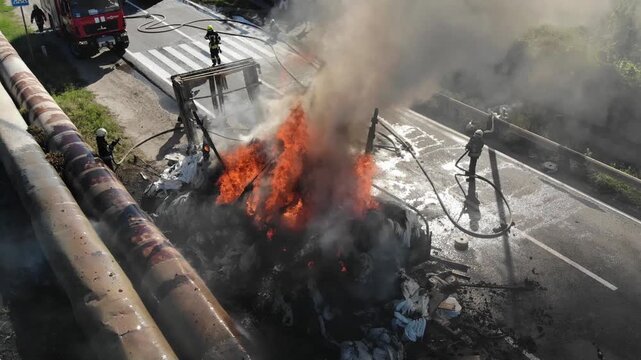 Critical situation: Firefighters are in the midst of a large fire near a roadway and transport infrastructure. Drone footage shows bright flames and smoke filling the road.