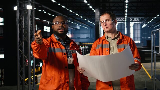 Diverse engineers handling assembly workflow with industrial plans, standing near steel machinery and fabrication stations that represent heavy industry development. Welding sparks.
