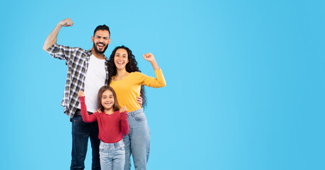 A smiling family stands proudly together, raising their arms in celebration. They are in casual outfits, exuding joy against a vibrant blue backdrop. © Prostock-studio