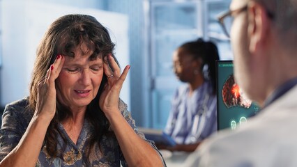 Senior woman at checkup deals with a painful headache and asks for advice at medical facility, recommending a treatment to relieve pain and ease the inflammation. Health insurance. Camera B.