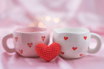 Two cups with hearts and knitted heart on light pink background with bokeh background